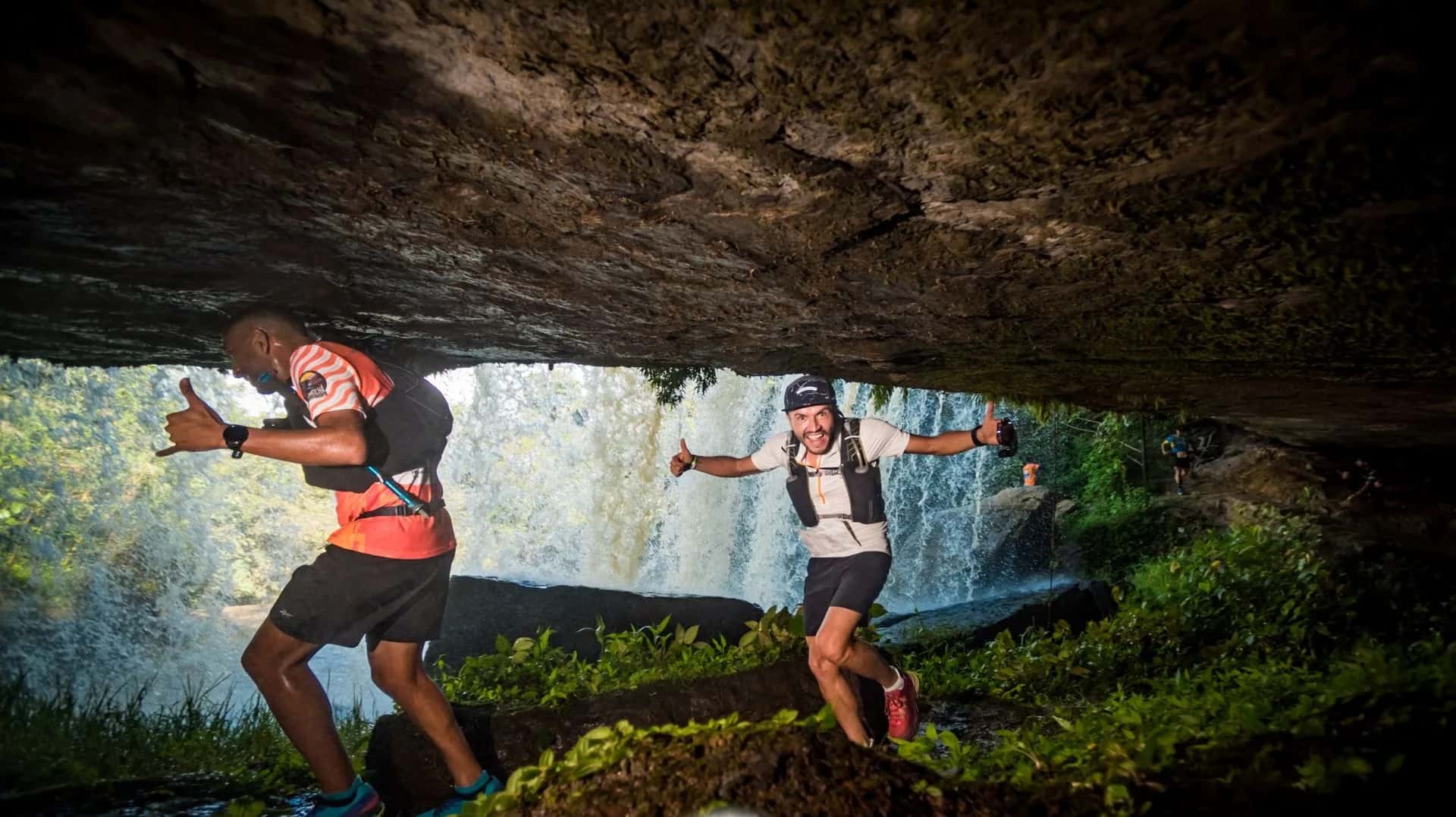 Two runners passing behind a waterfall, under a rock overhang, on the 2023 course