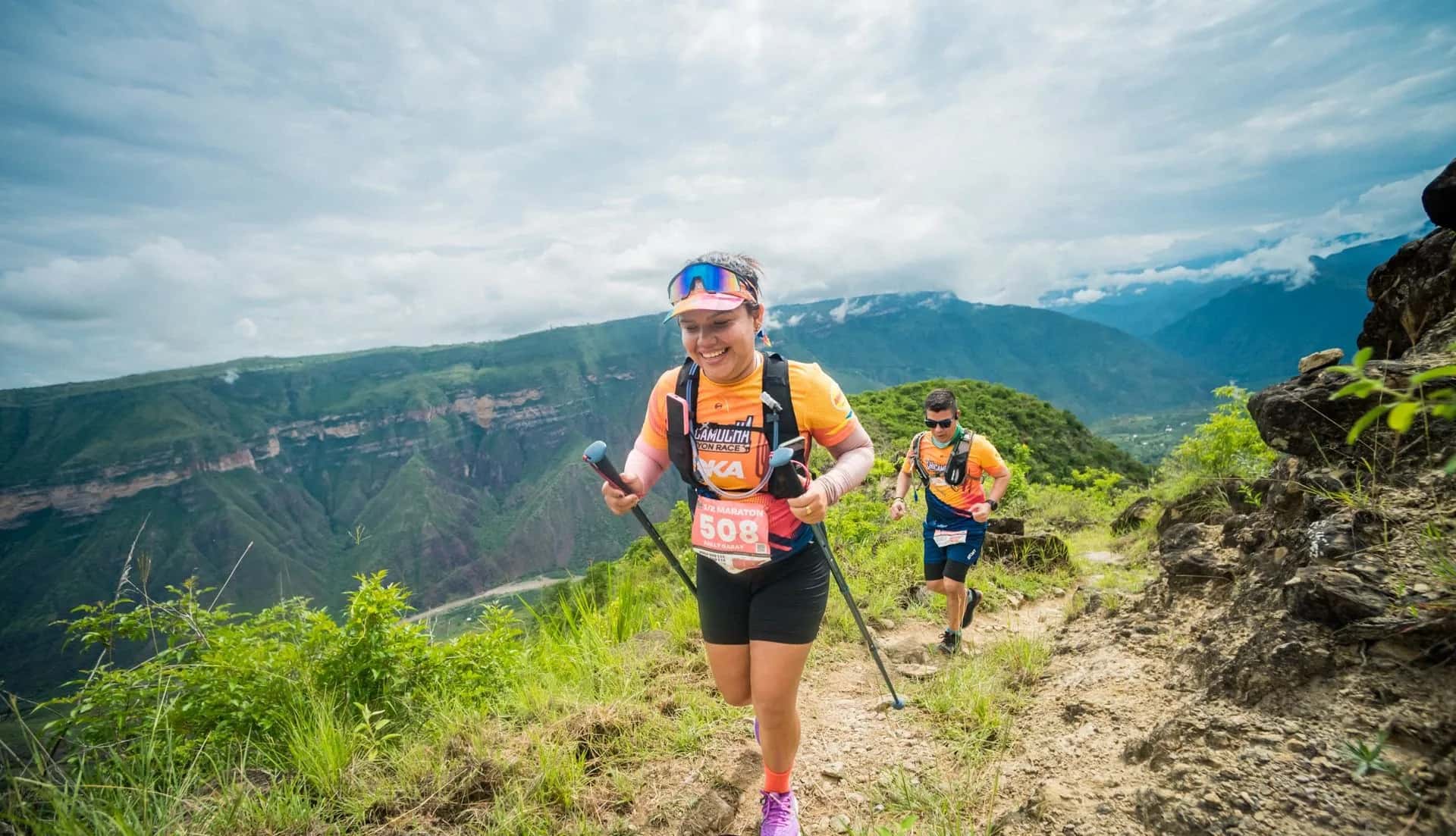 Smiling runner with poles on a hillside trail, followed by another runner, with the canyon and river behind