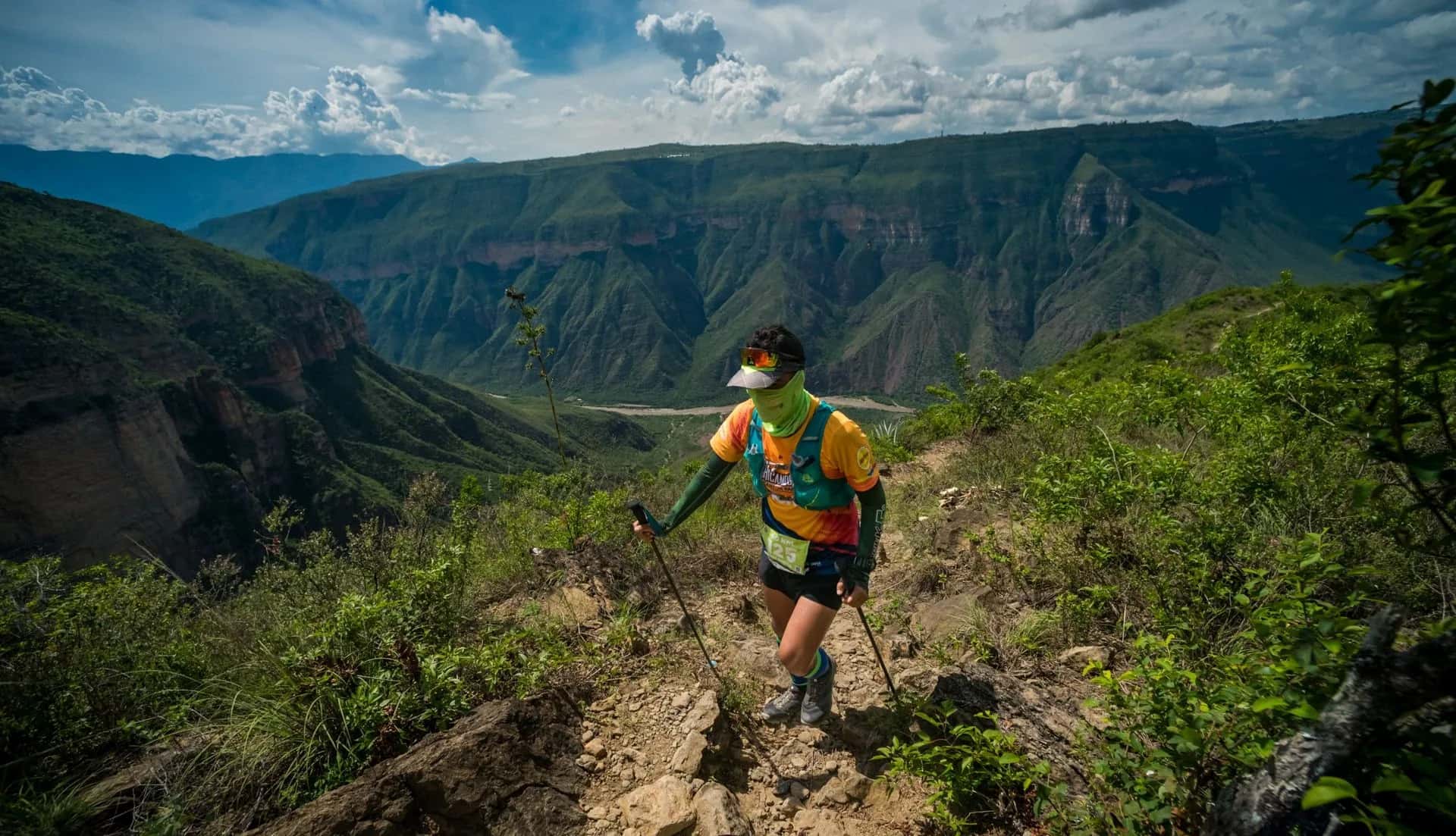 Runner with poles climbing a dirt trail, with the Chicamocha Canyon walls and river behind