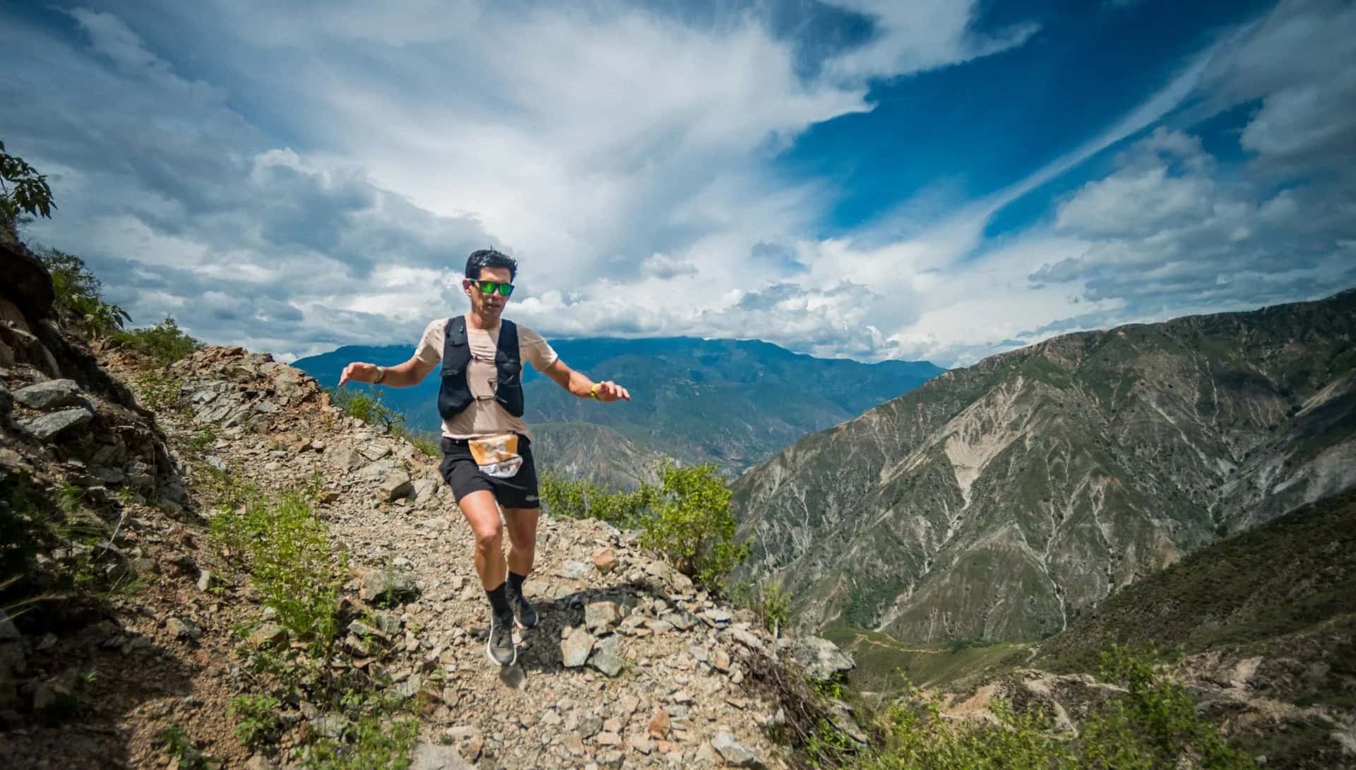 Runner with a hydration vest descends a rocky ridge, with the canyon and a cloudy sky behind