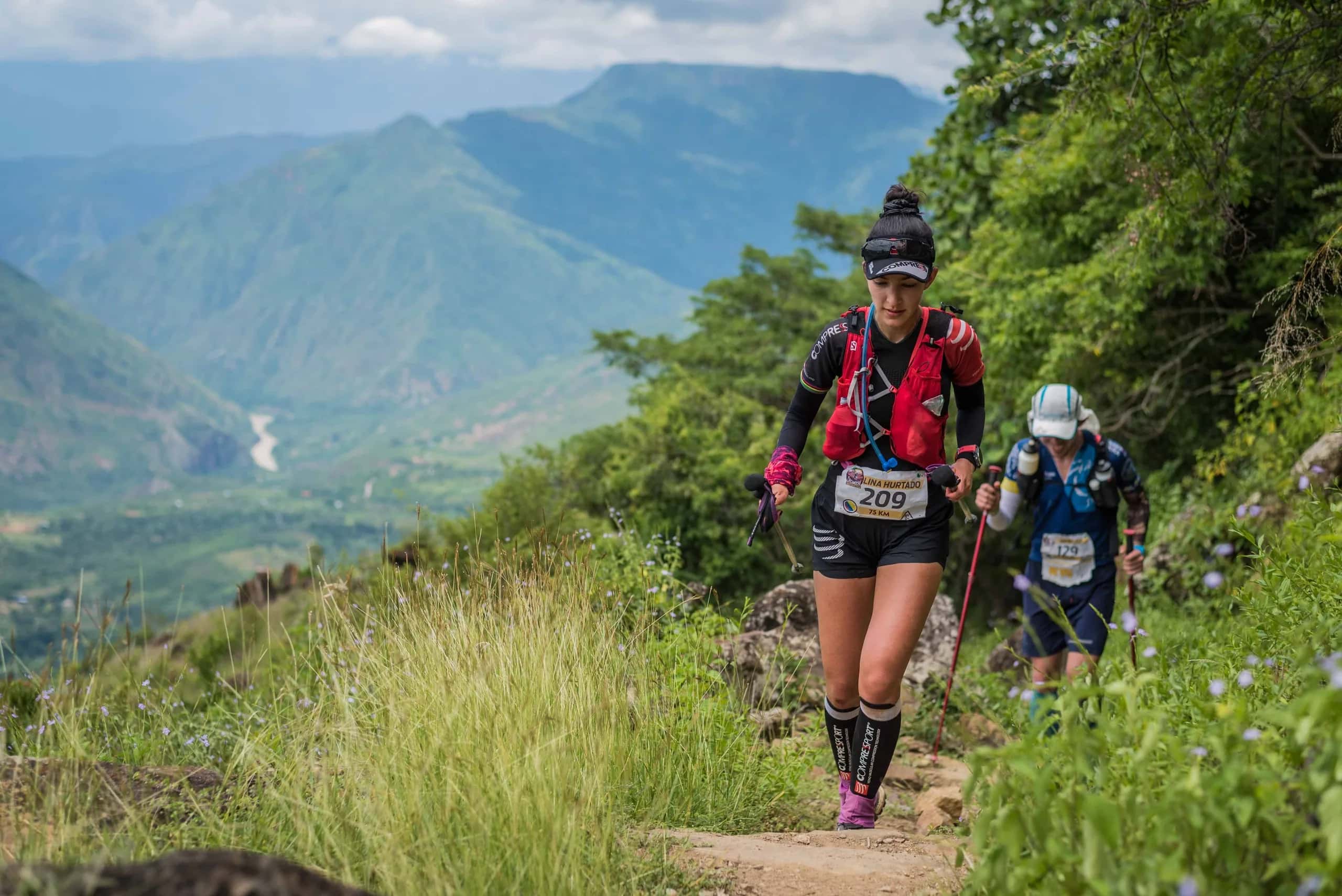 Two runners with poles on a canyon trail, with the valley and river far below