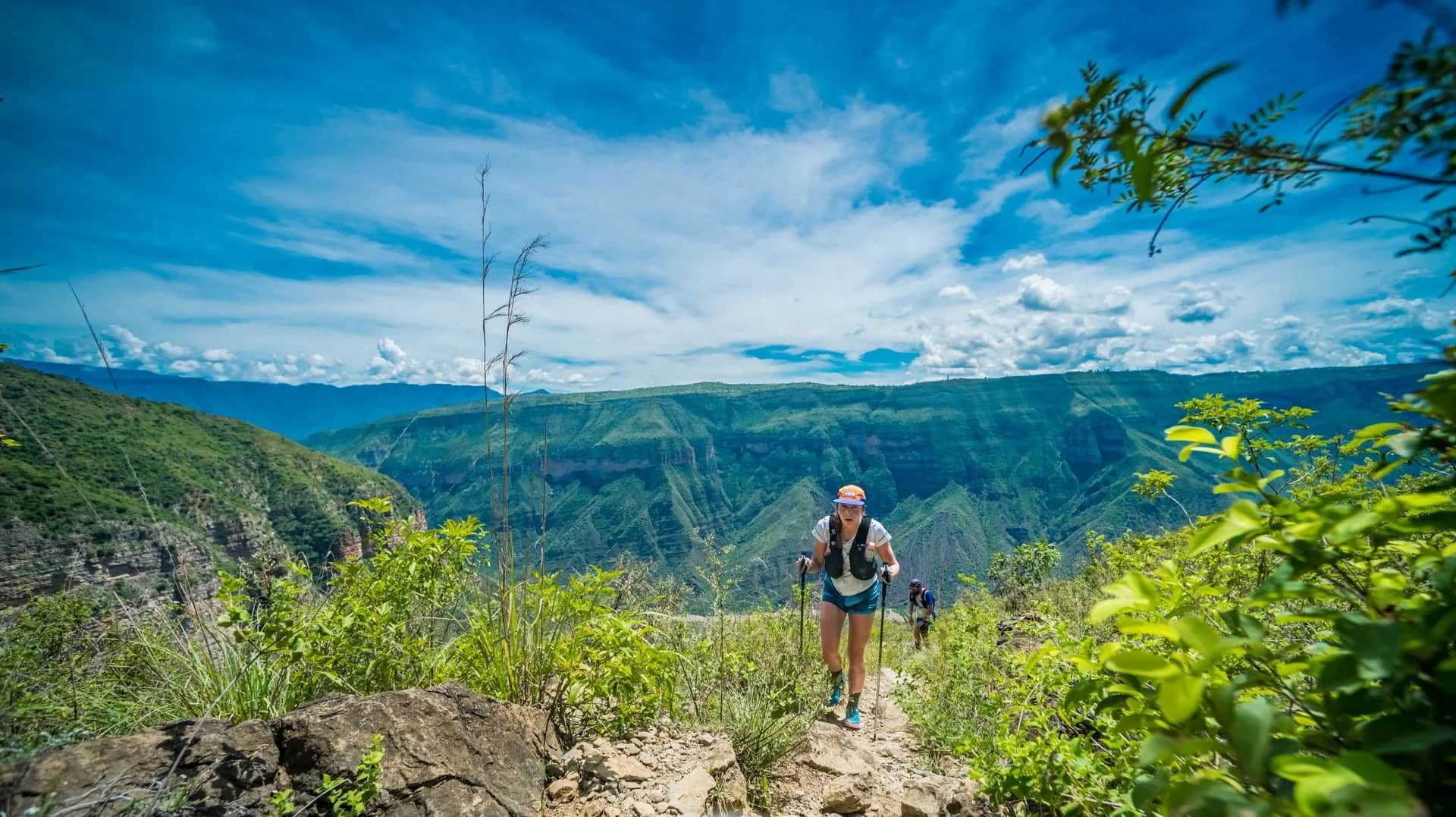 Runners moving along a rocky trail on the canyon rim under a blue sky with clouds
