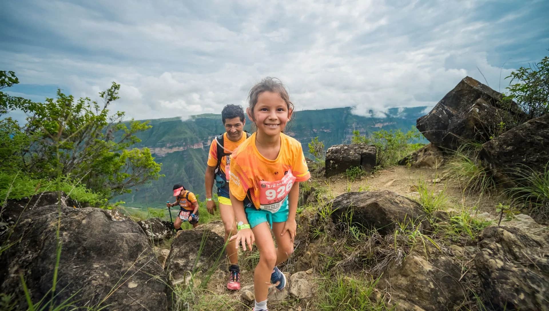 Young girl smiles as she climbs a rocky trail, followed by other runners