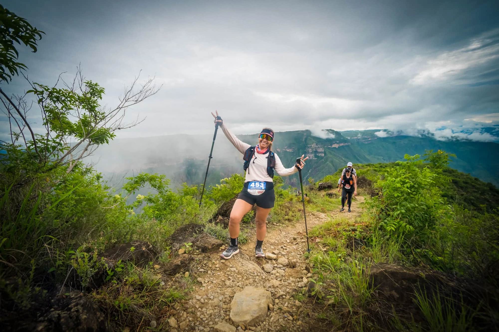 Smiling runner with poles waving on a trail through the vegetation, clouds over the canyon