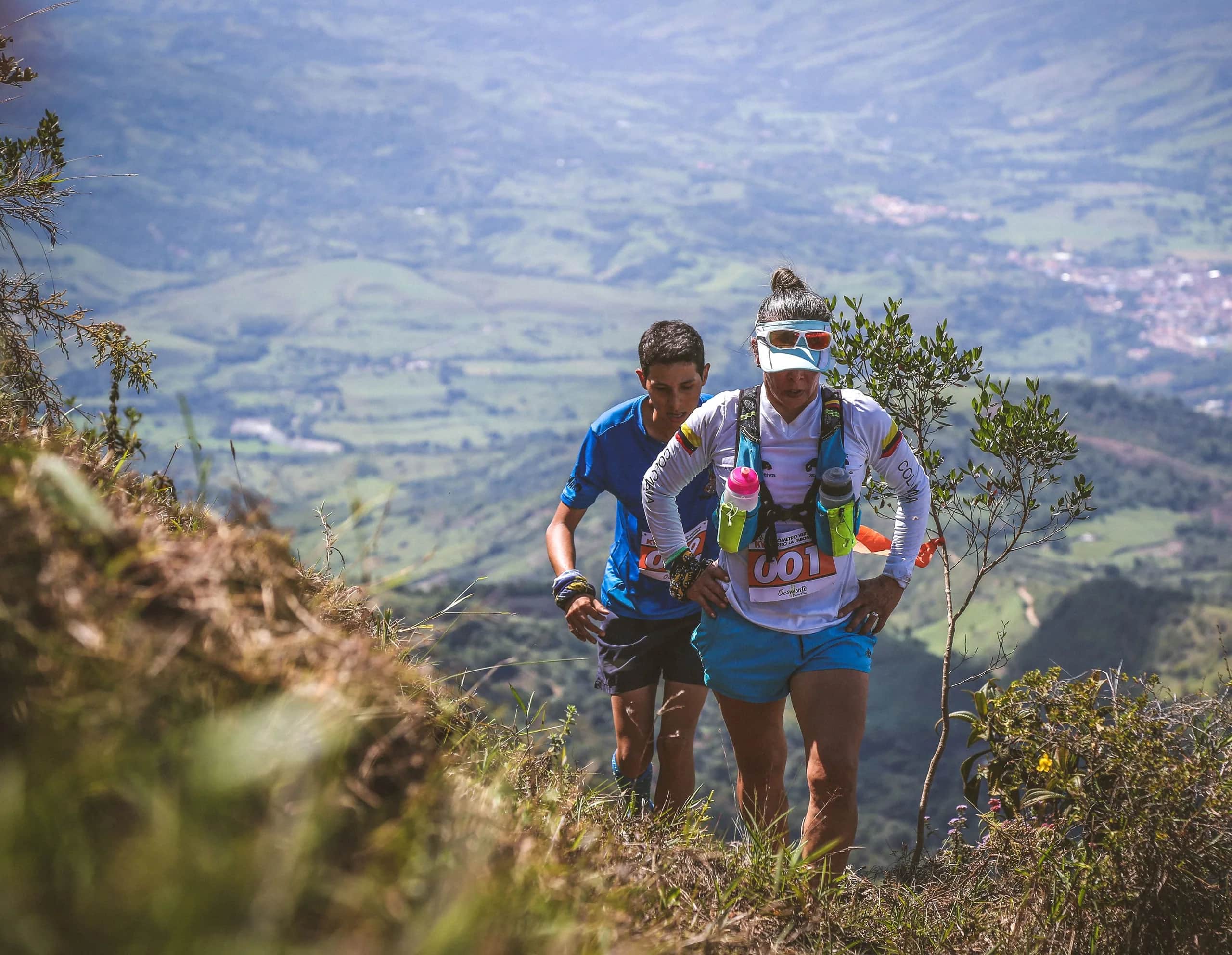 A woman and a man climbing the hillside together, with the valley and a town below