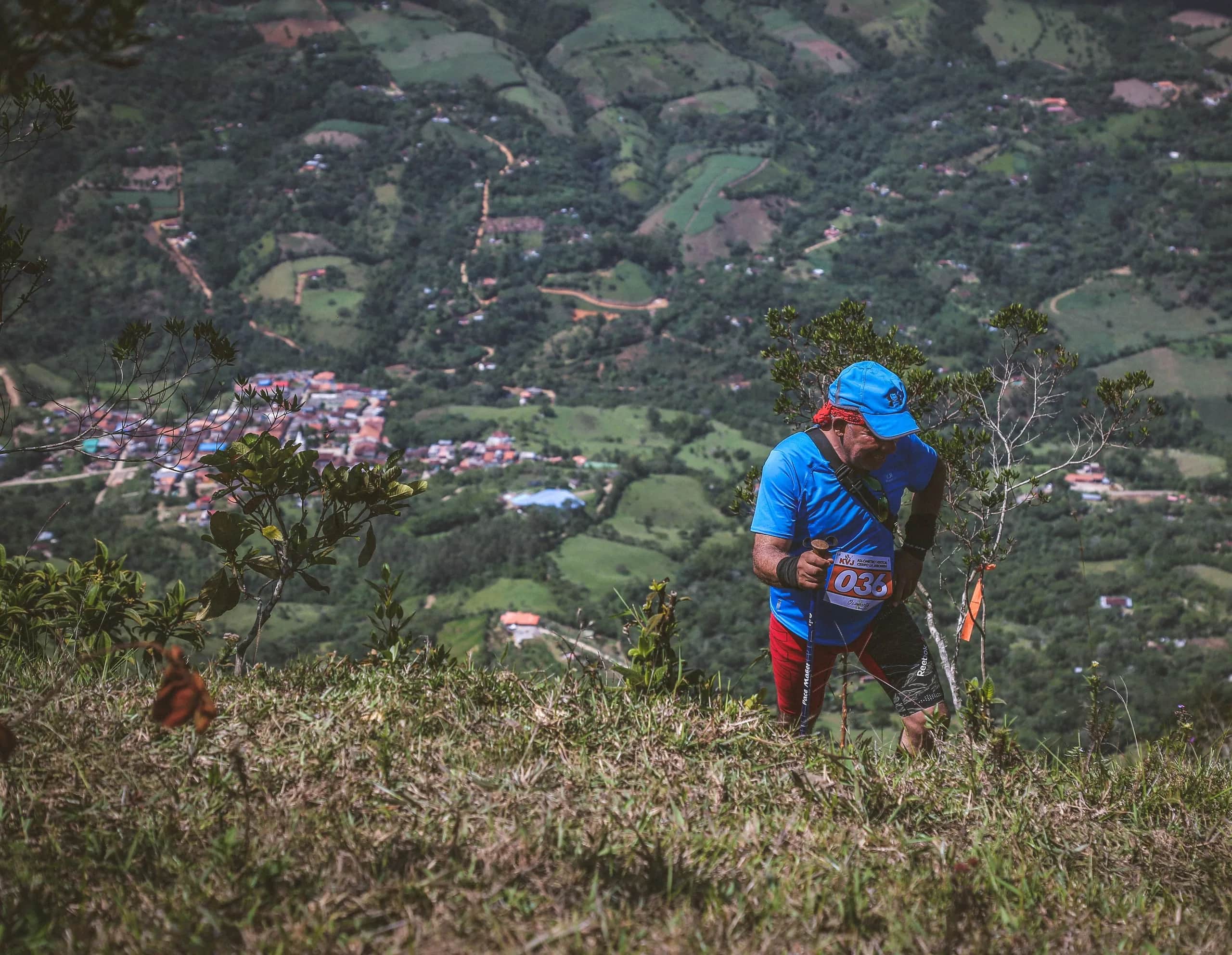 Runner in a blue cap climbing through shrubs, with Ocamonte and the valley farmland below