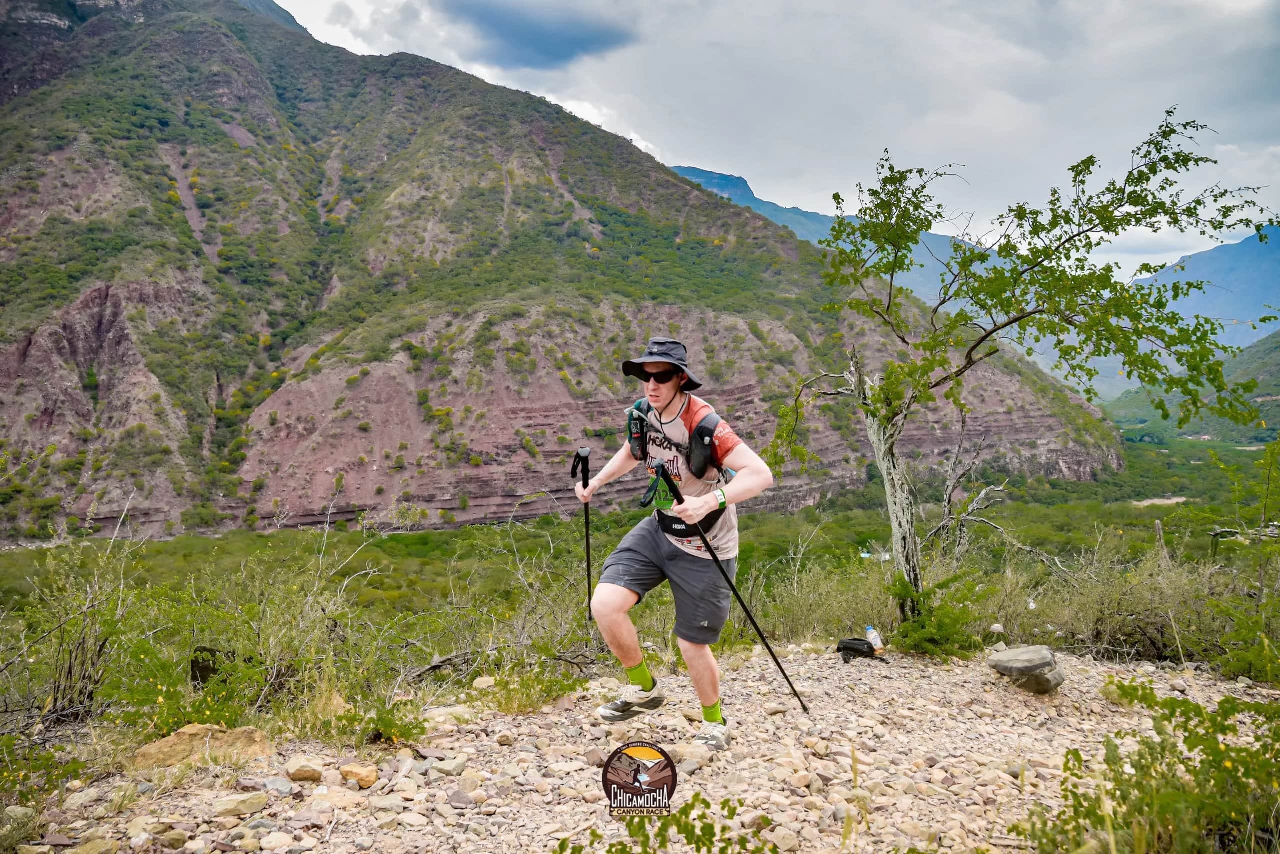 Corredor con sombrero y bastones avanza entre arbustos, con el cañón y las montañas al fondo