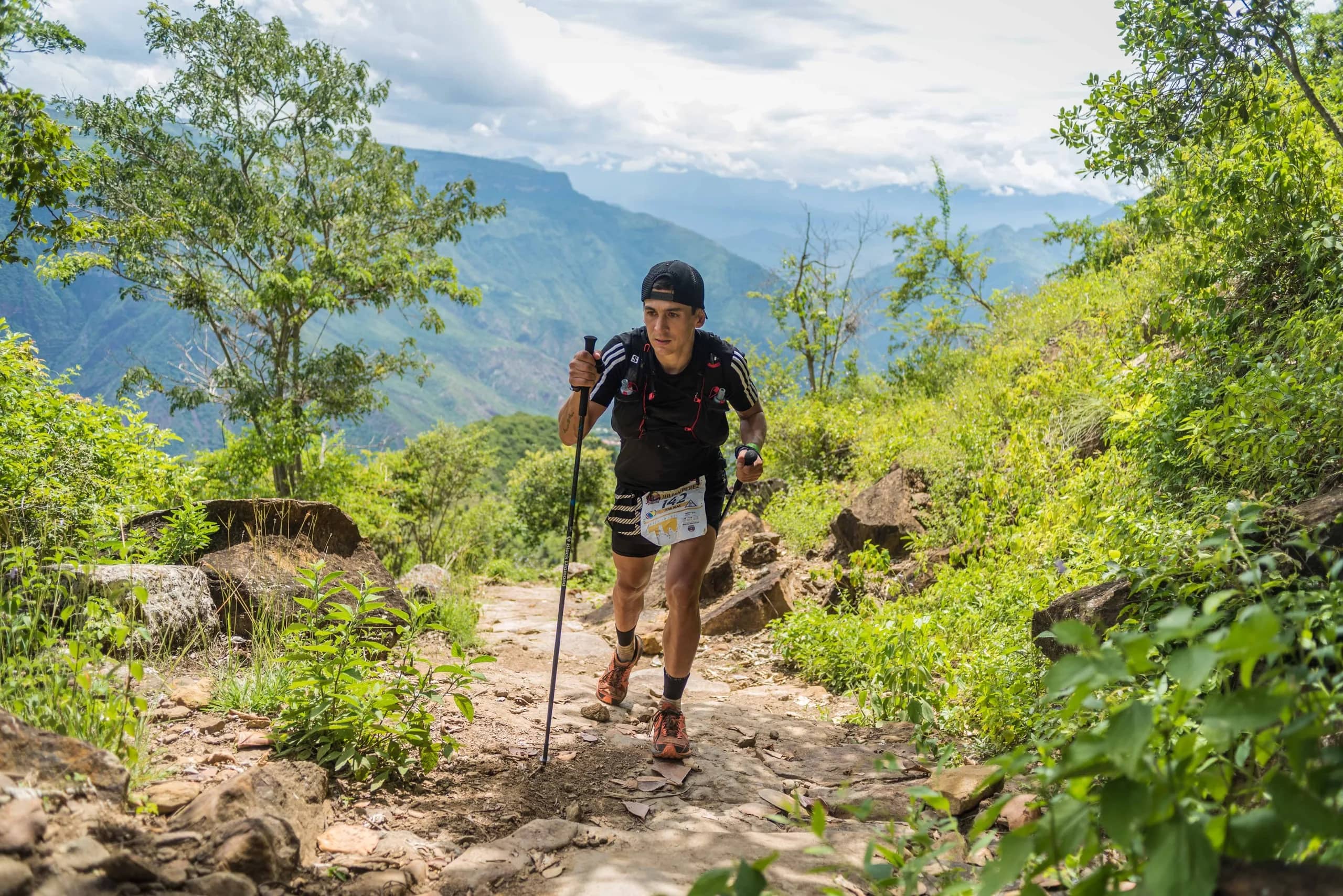 Runner with poles climbing a stony path through the vegetation, with the canyon behind