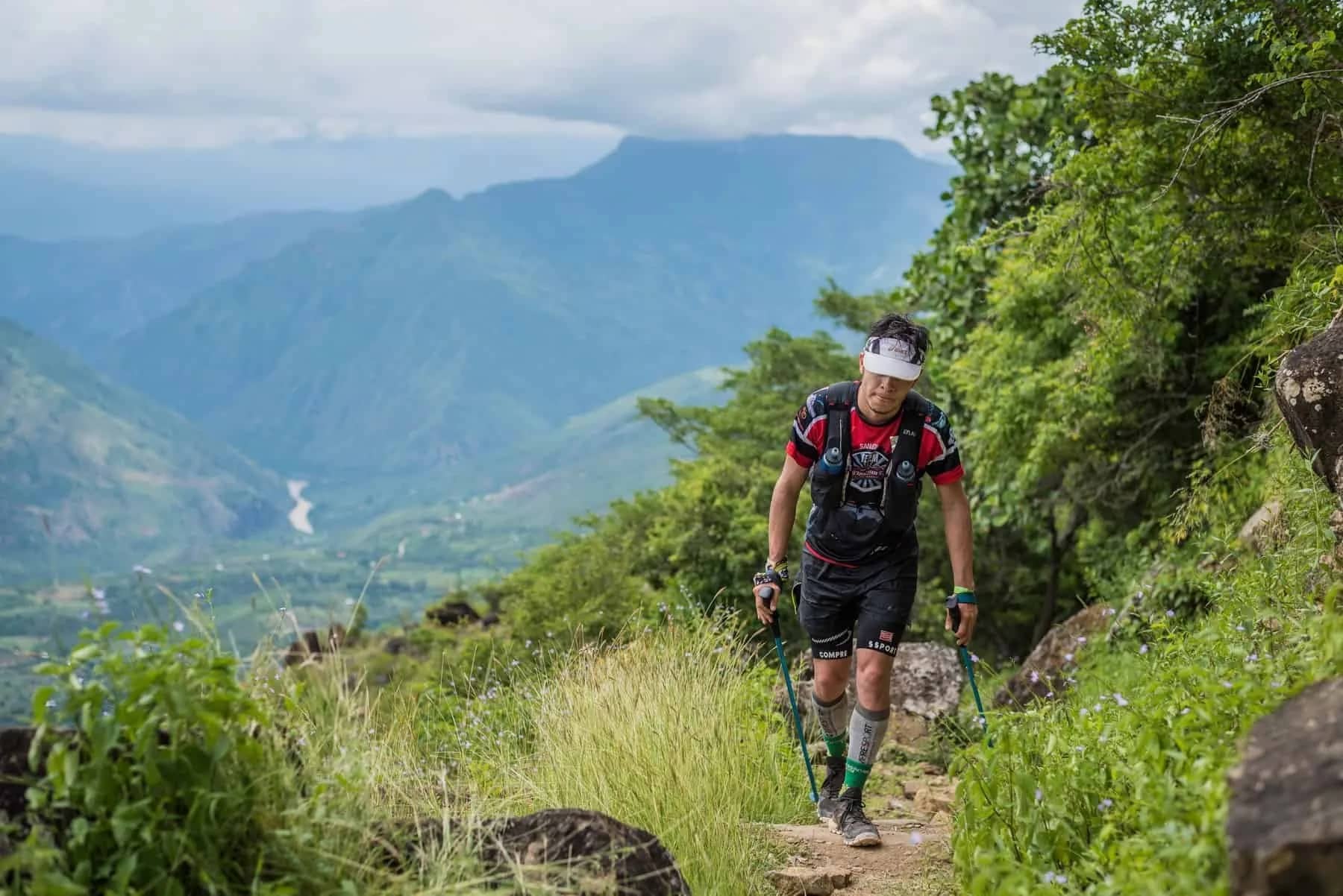 Runner with poles climbing a mountain trail, with the river winding along the valley floor below