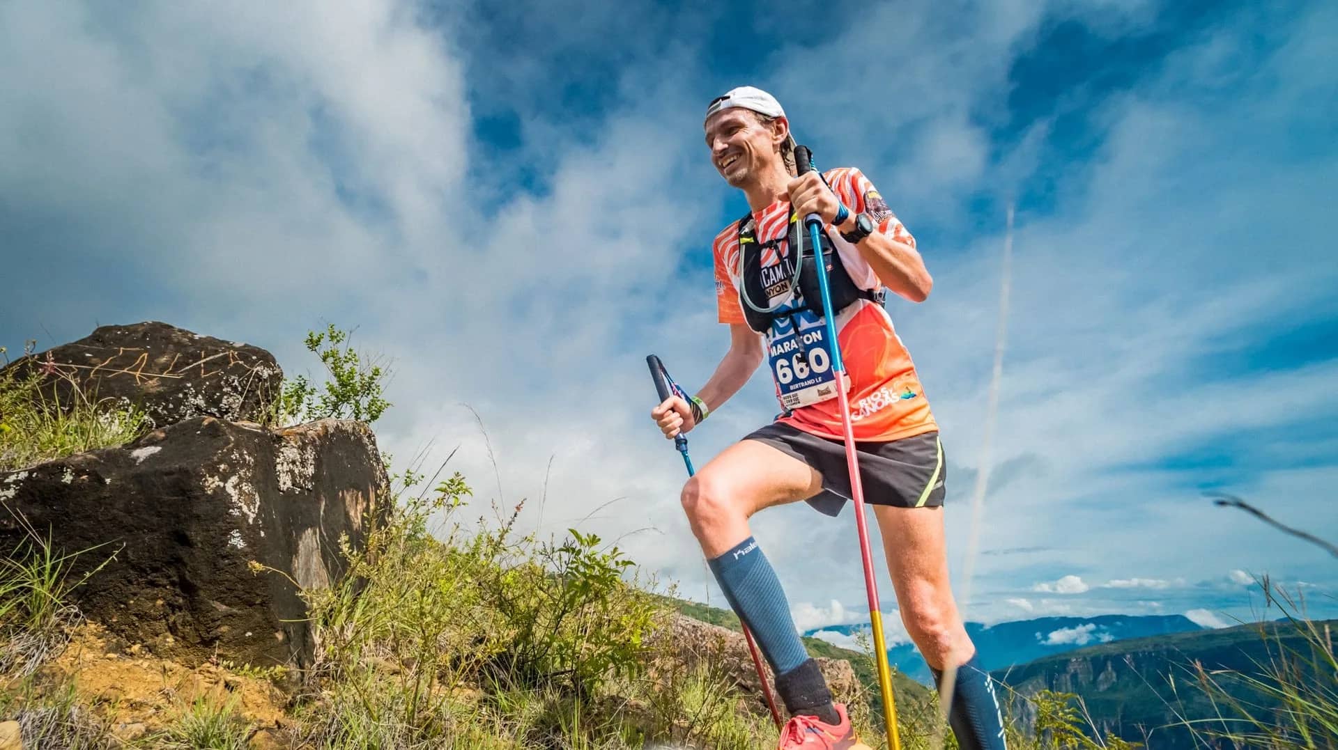 Marathon runner with poles climbing a rocky slope under a blue sky with clouds
