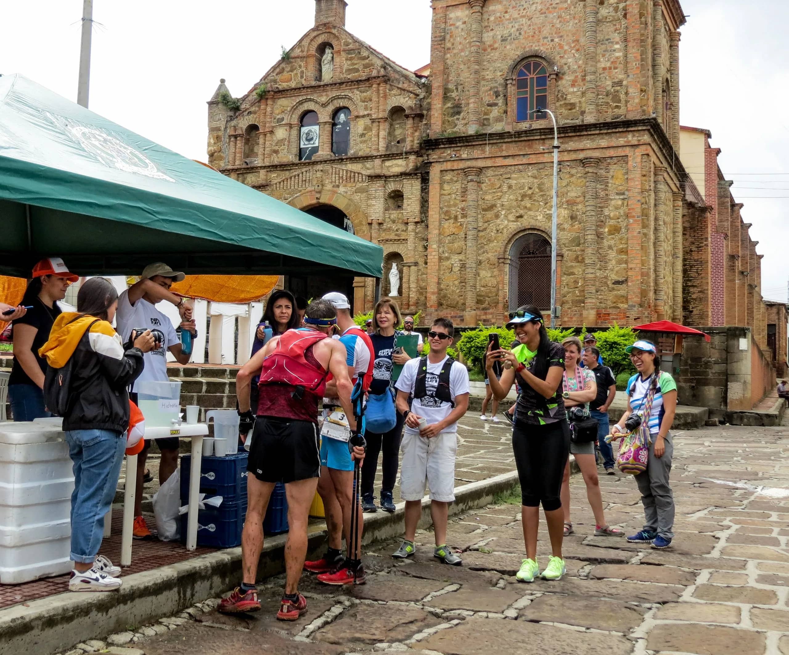 Corredores y público en un puesto de hidratación frente a una iglesia de piedra en la plaza de un municipio del recorrido