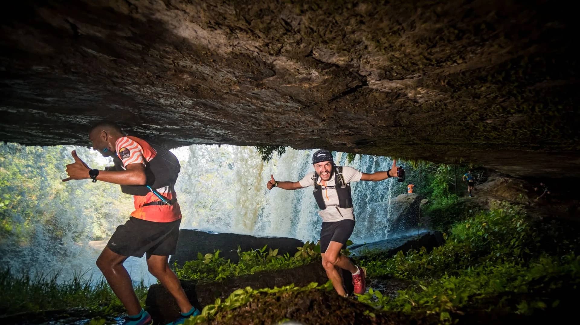Two runners passing behind a waterfall, under a rock overhang, on the 2023 course