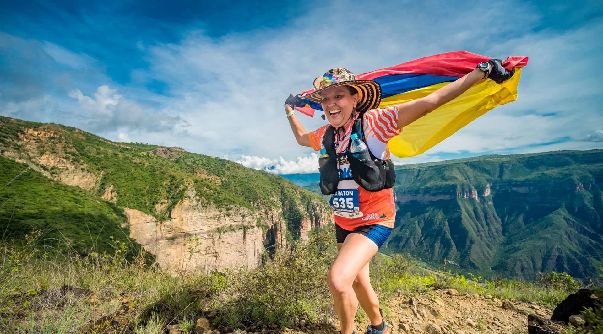 Runner in a hat with the Colombian flag over her shoulders celebrating on the canyon rim