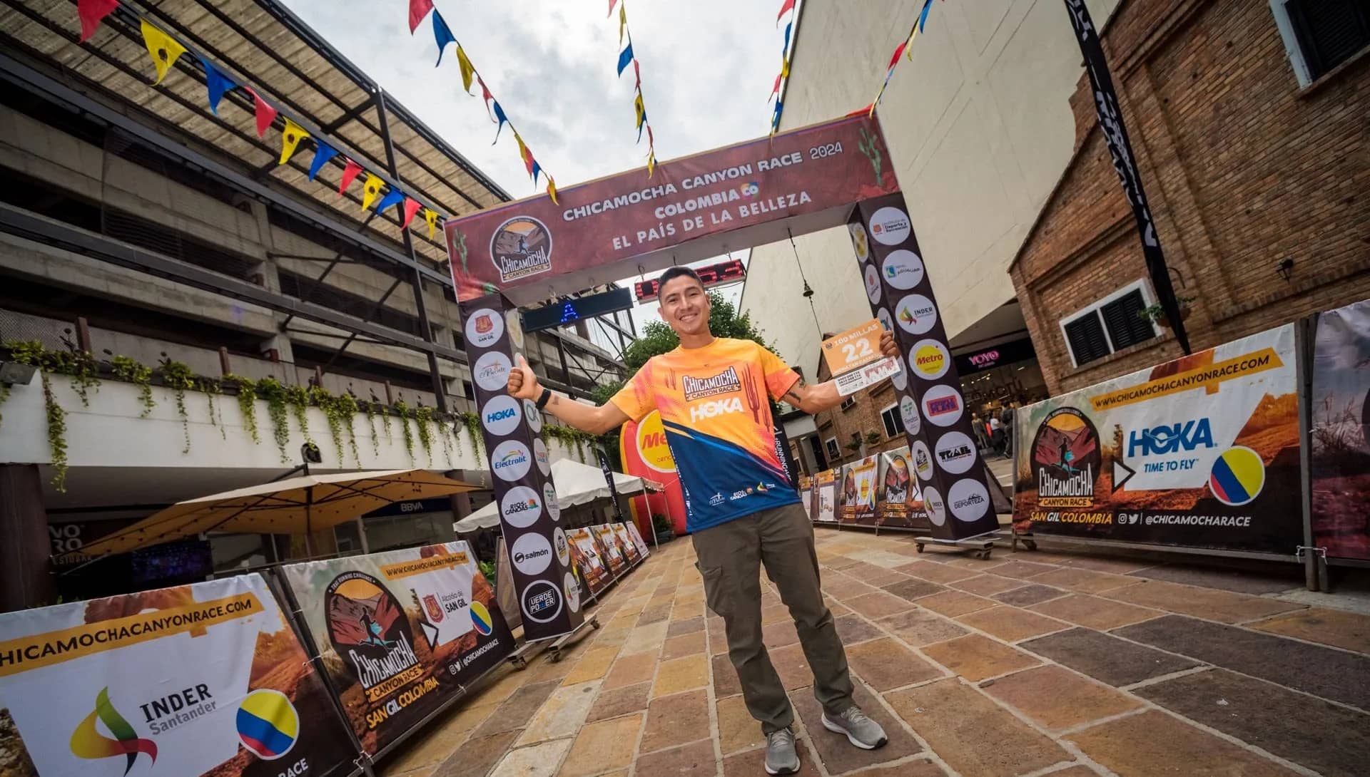 Runner poses in the official race shirt, holding his bib, under the start and finish gantry