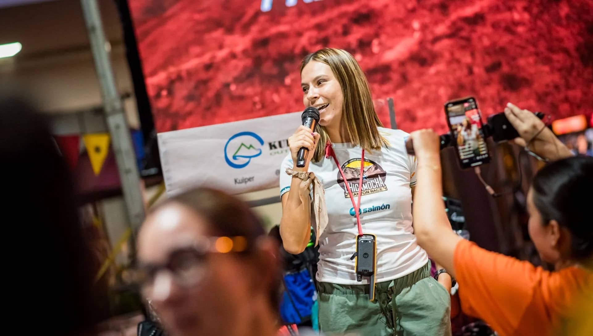 Host with a microphone at a runner expo stand, surrounded by people taking photos
