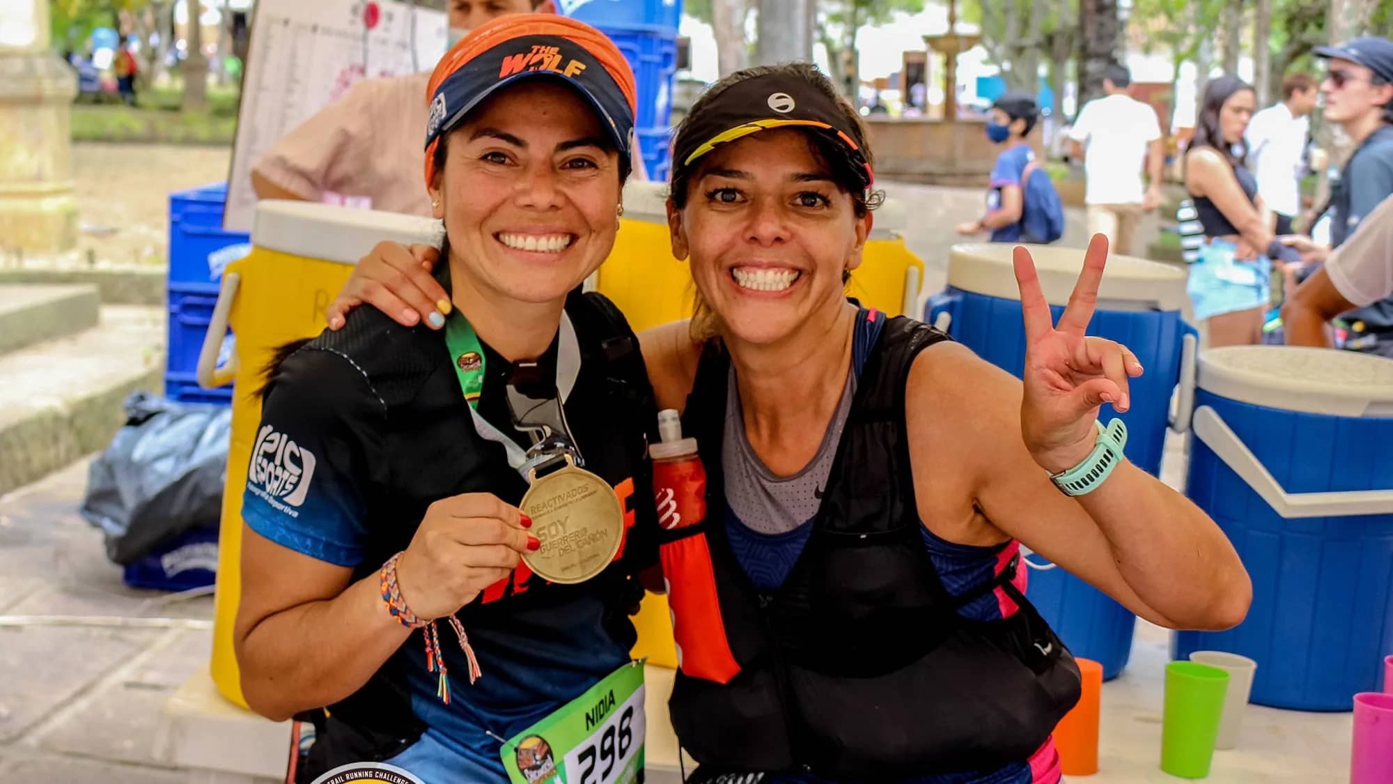 Two smiling runners showing their finisher medal next to a hydration table