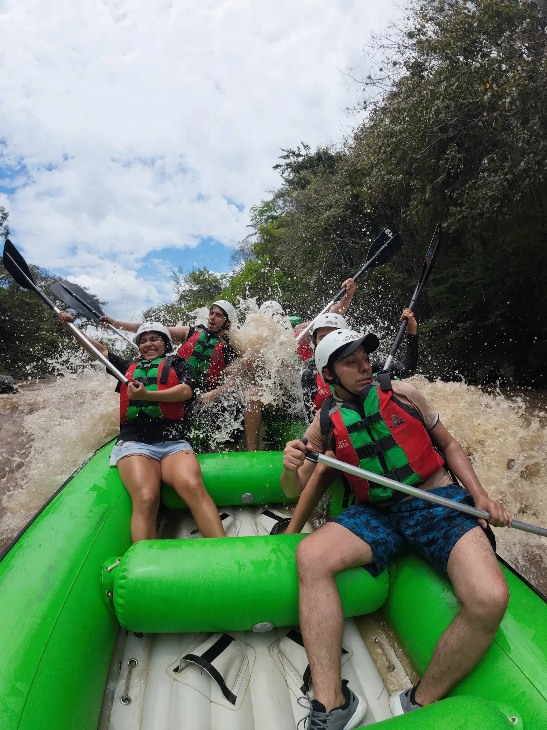 Group in helmets and life jackets crashing through a rapid in a green raft, paddles up amid the spray