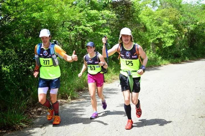 Three runners wearing bibs moving along a road lined with vegetation, smiling
