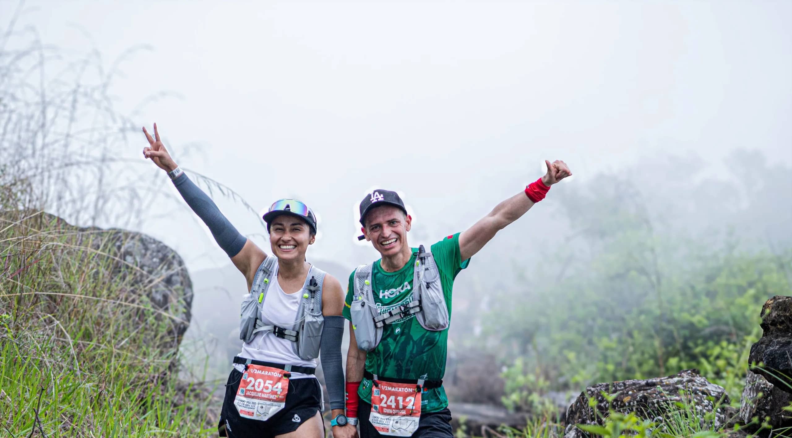 Two runners with race bibs celebrate with arms raised on a misty trail